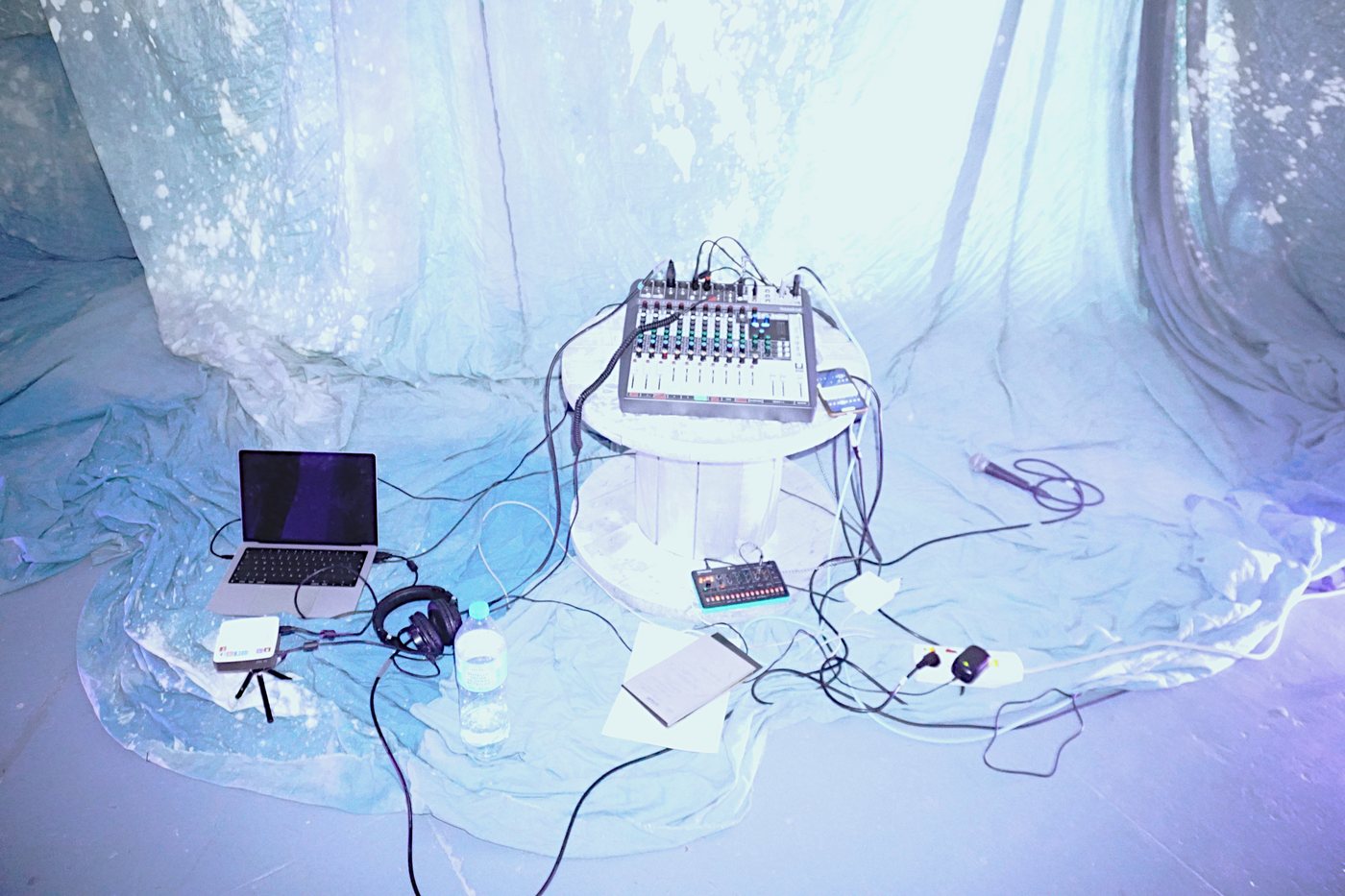 A sound setup on a table among hanging painted fabrics, lit in blue.