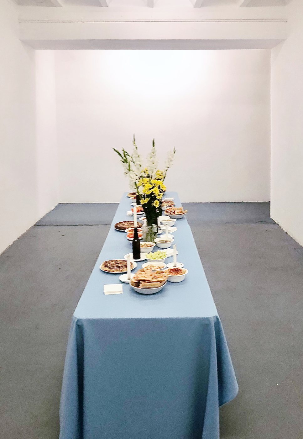 A long table with a blue cloth, flowers, bread and fruit, in a white gallery.
