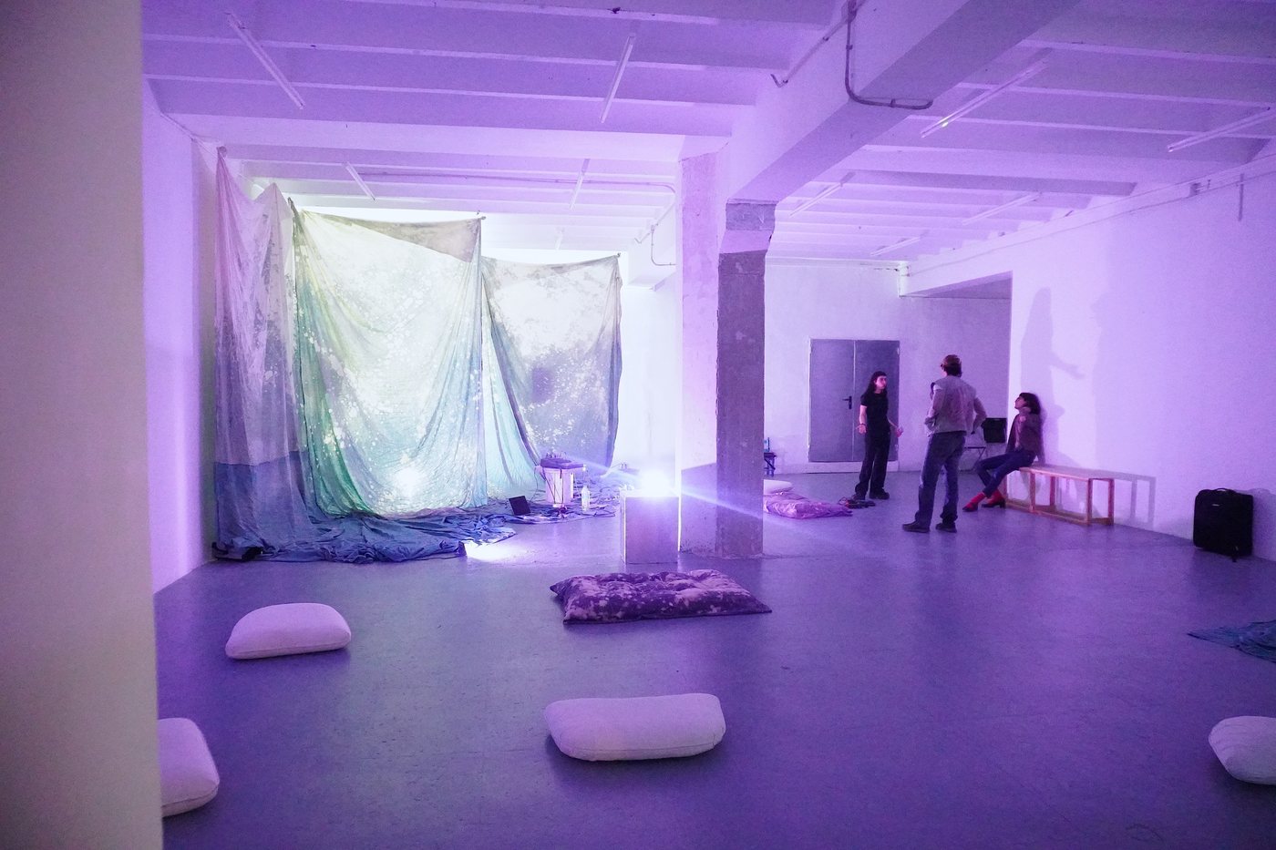 A gallery lit in blue, with hanging painted fabrics, cushions on the floor and people talking, at El Pati de Malpaís.