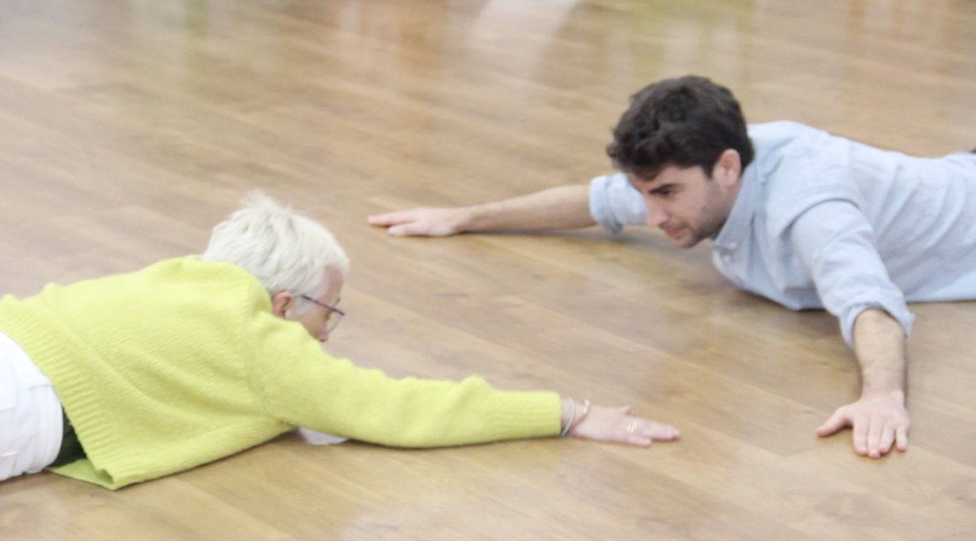 Two people lying on a wooden floor, reaching towards each other, in La sombra que deja el hilo.