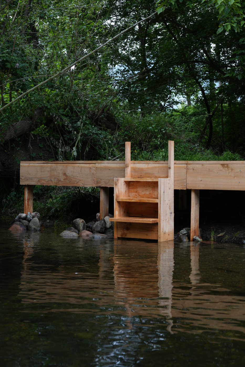 A wooden jetty with steps over a river, under green trees, reflected in the water.