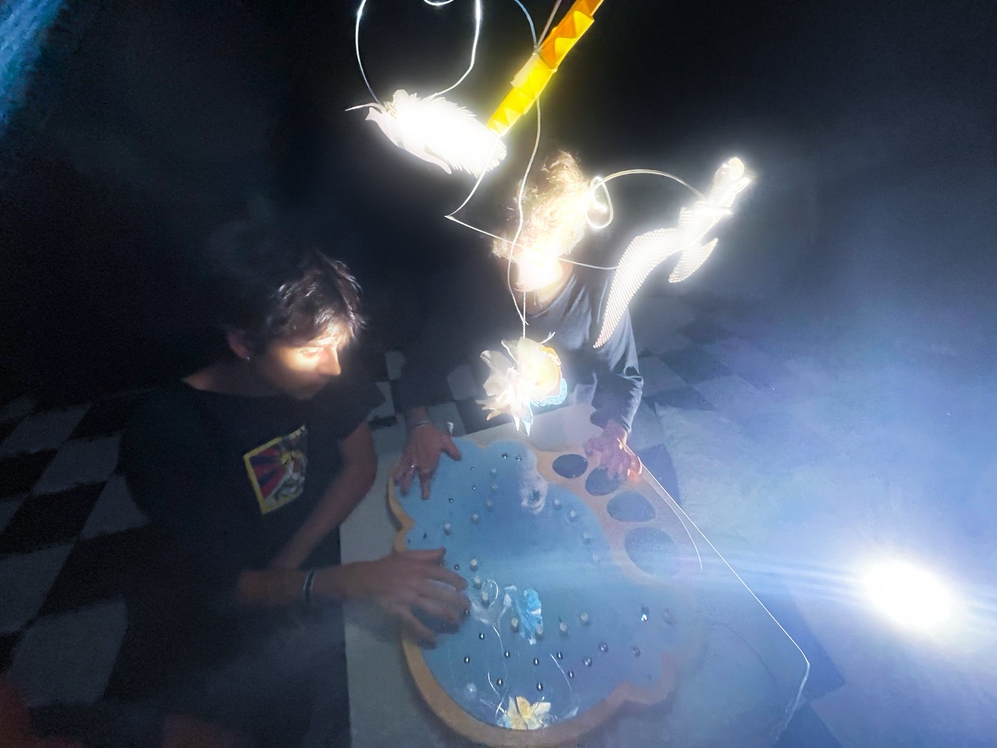 Two people playing Núvol, a cloud-shaped instrument lit by glowing figures hanging above it, in the dark.